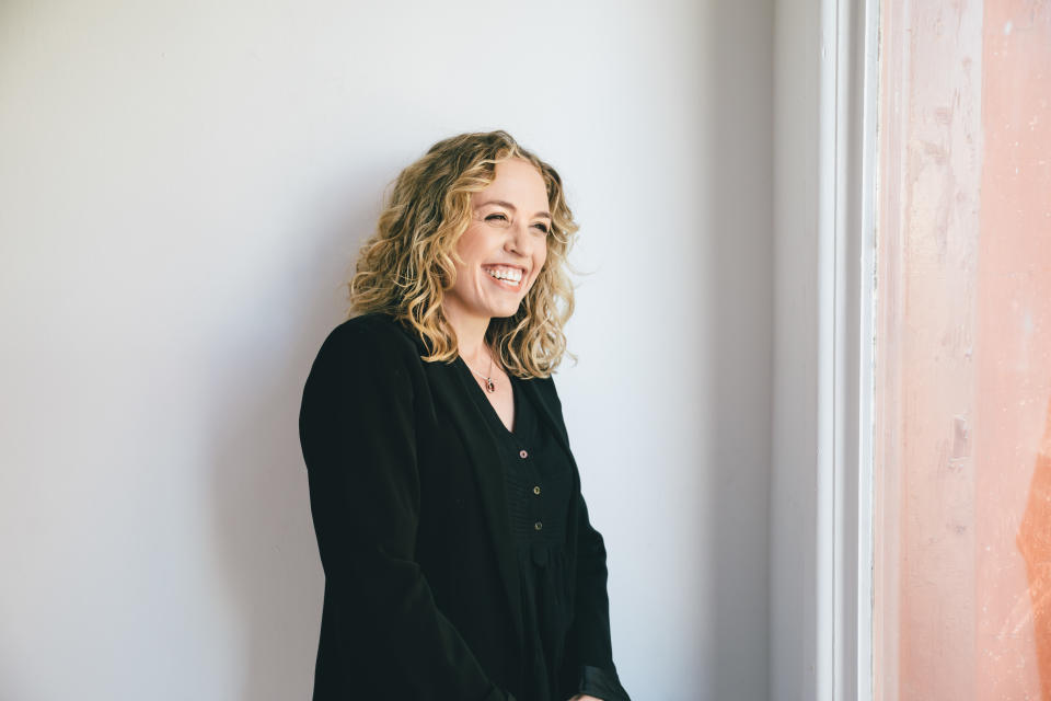 CEO Kristy Chong laughs in three-quarter profile against a white wall, while wearing a black garment. She has blond streaked curly hair worn loose.