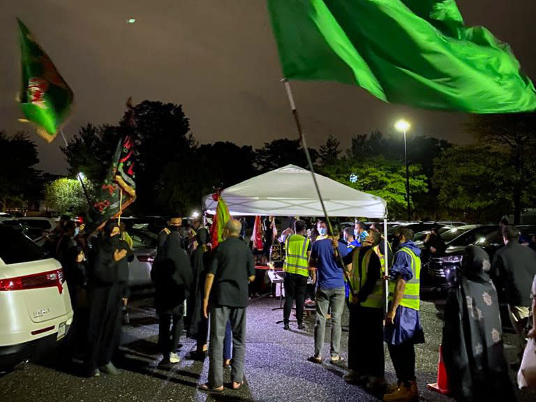 In a photo provided by Fatima Mukhi-Siwji, people gather with flags in a parking lot for a drive-in commemoration of the seventh-century death of Imam Hussein, Thursday, Aug. 27, 2020, in Hicksville, N.Y. Shiite Muslims are marking the mourning period in the shadow of the coronavirus. (Fatima Mukhi-Siwji via AP)(Fatima Mukhi-Siwji via AP)