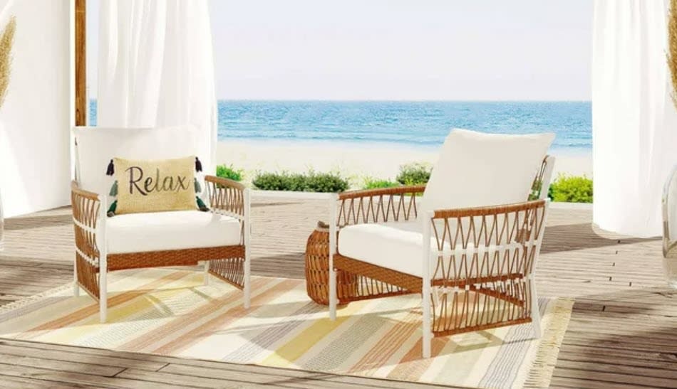 Two woven chairs with cushions on a deck by the beach, one with a "Relax" pillow. No persons present