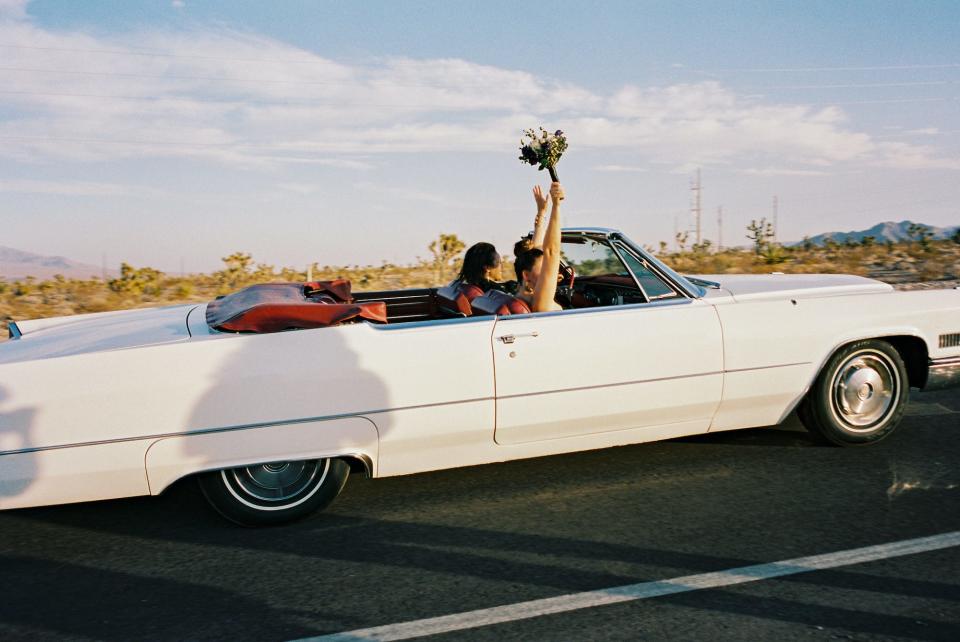 A bride holds her bouquet up as they drive in a convertible on their wedding day.