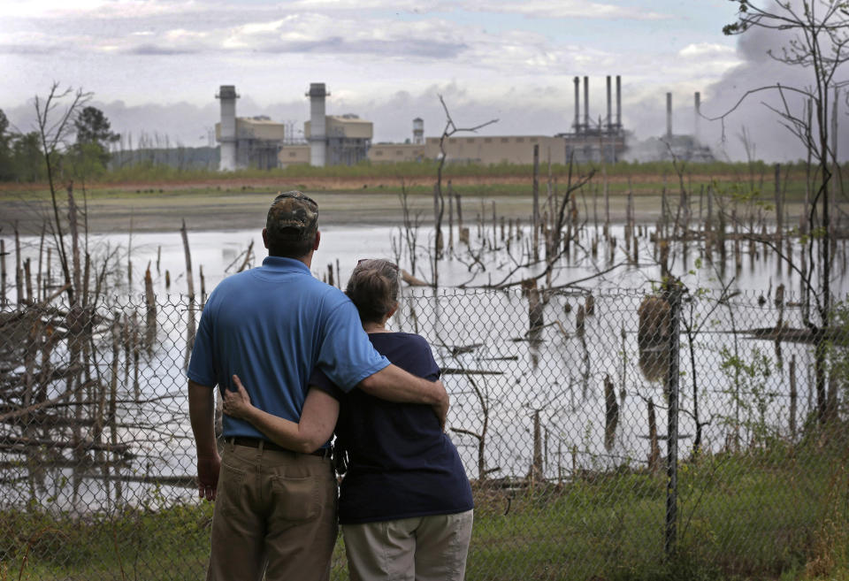 FILE - In this April 25, 2014, file photo, Bryant Gobble, left, embraces his wife, Sherry Gobble, right, as they look from their yard across an ash pond full of dead trees toward Duke Energy's Buck Steam Station in Dukeville, N.C. The federal, North Carolina and Virginia governments want a judge to declare Charlotte-based Duke Energy liable for environmental damage from a leak five years ago that left miles of a river shared by the two states coated in hazardous coal ash. Government lawyers on Thursday, July 18, 2019 sought to have Duke Energy declared responsible for harming fish, birds, amphibians and the bottom of the Dan River. (AP Photo/Chuck Burton, File)