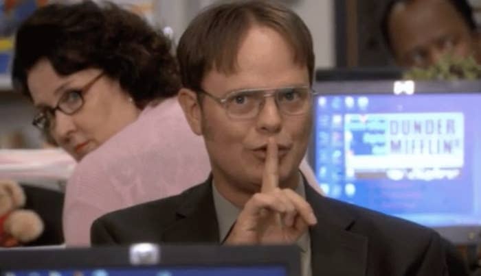 Dwight from "The Office" gestures for silence with a finger over his lips, Phyllis in background. They're in an office setting