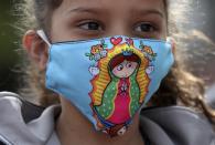 A Venezuelan migrant girl, wearing a protective face mask with a graphic version of the Virgin of Guadalupe, waits for a bus to travel to the border, after breaking camp in Bogota, Colombia, Thursday, July 2, 2020. Facing no work due to the COVID-19-related economic shutdown, hundreds of Venezuelan migrants are returning to their country. (AP Photo/Fernando Vergara)