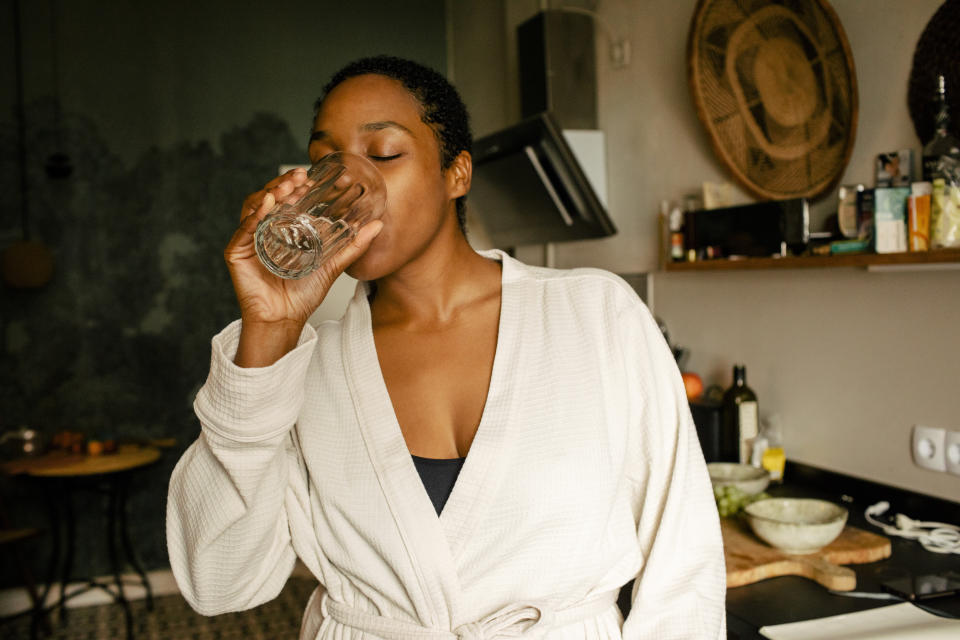 Person wearing a bathrobe standing in a kitchen and drinking water from a glass