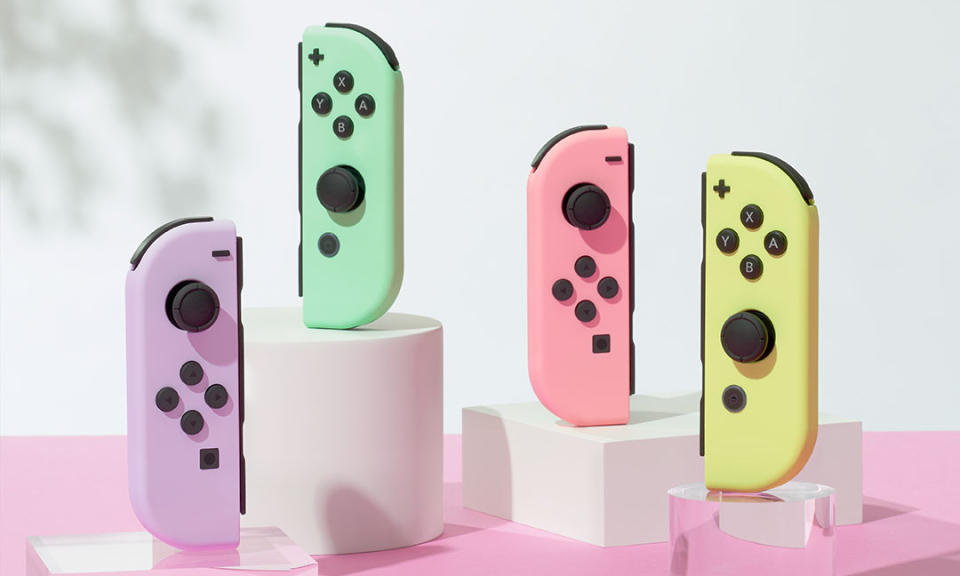Marketing photo of two pairs of pastel colored Nintendo Switch Joy-Con controllers.  From left to right: purple (left controller), green (right controller), pink (left controller), and yellow (right controller).  They sit upright on stands on a pink table with a white background.