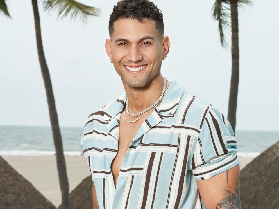 Will Urena poses in a striped shirt and tan shorts in front of a pool with a beach in the background.
