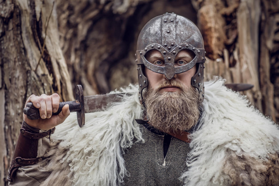 A bearded person dressed in Viking attire, wearing a metal helmet and fur cloak, holding a sword over their shoulder, in a forest setting