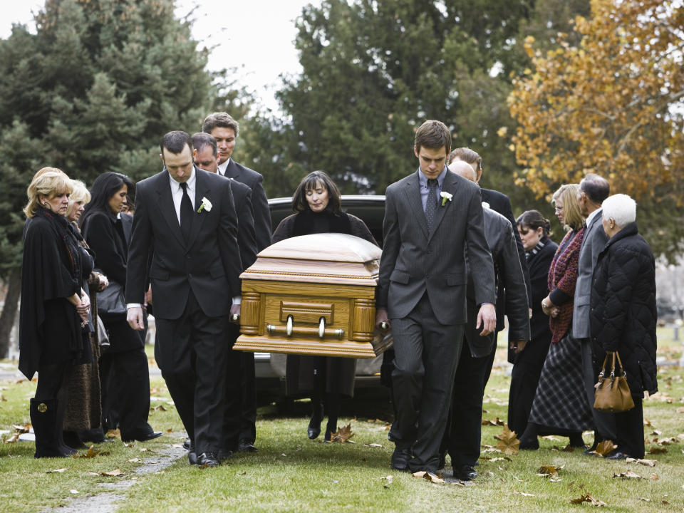 A group of people dressed formally in suits and dresses are carrying a casket towards a hearse at a funeral with onlookers standing nearby