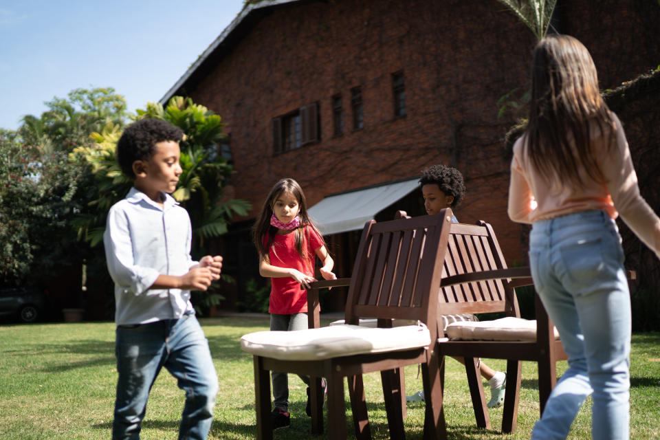 Four kids walk around a group of three chairs in a backyard.