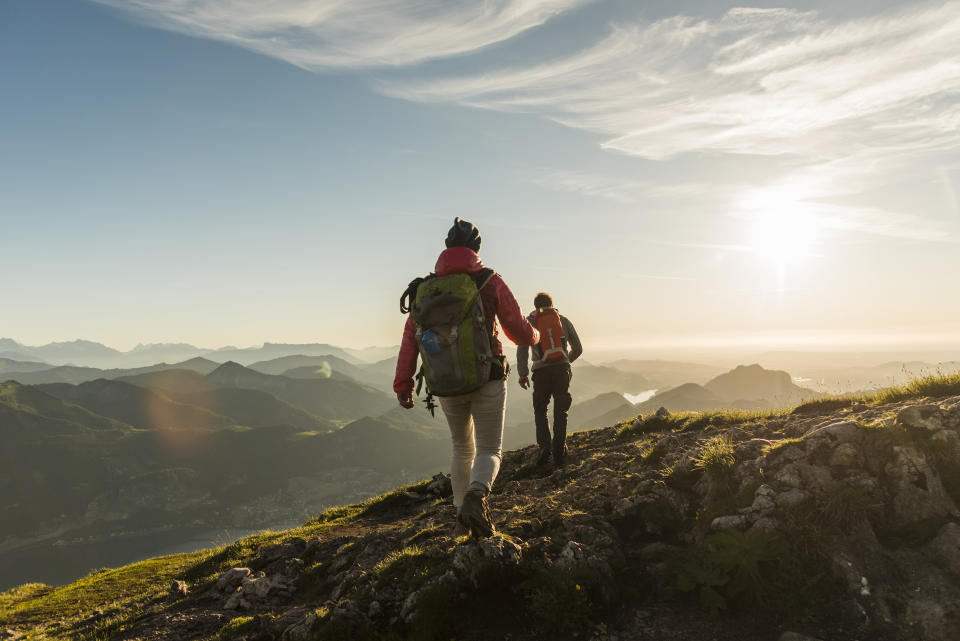 Two hikers on a mountainous trail during sunrise, wearing backpacks and outdoor gear, with a scenic landscape of mountains and valleys in the background