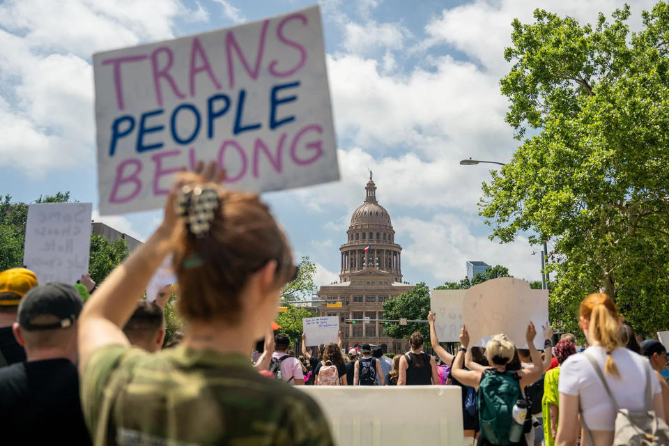 A protesters holds up a sign that reads "Trans People Belong" as the Texas State Capitol can be seen in the background.