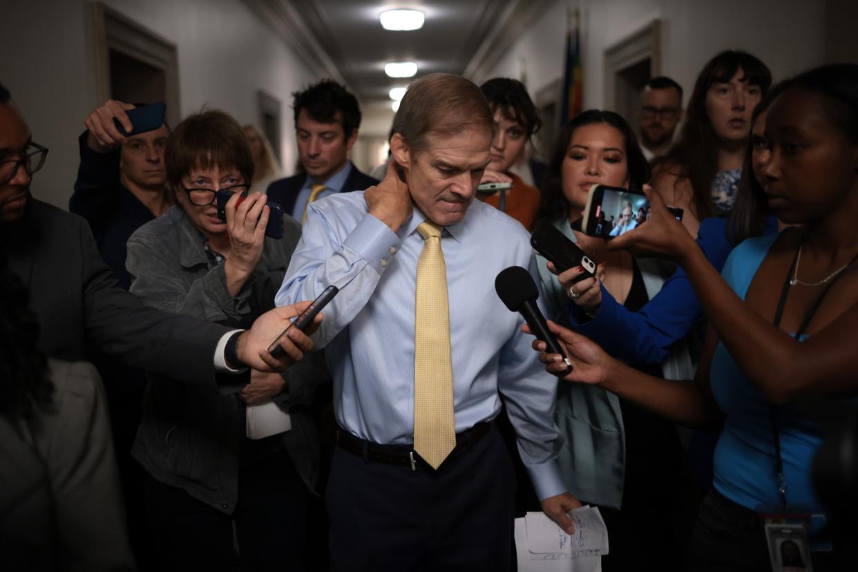 Rep. Jim Jordan, standing in a hallway surrounded by reporters holding out their microphones and voice recorders, rubs the back of his neck and looks downward.