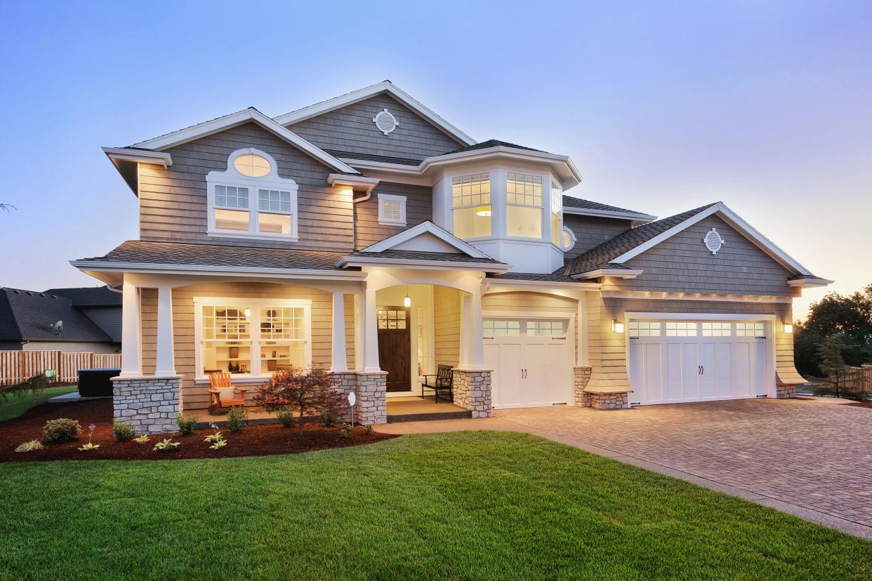 Front of large suburban home with landscaped lawn and a driveway, with lights on during early evening, clean on a summer day, with a dark blue sky