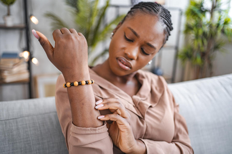 A woman sitting on a chair examines her elbow carefully, appearing to check for problems.  She wore a beige blouse and a beaded bracelet