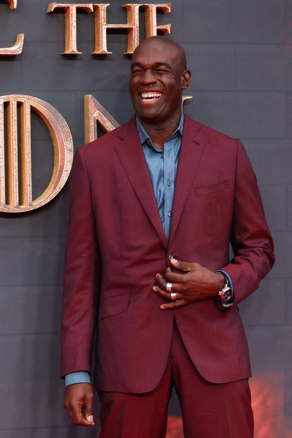 A black man wearing a dark red suit with a blue shirt underneath, smiling on a red carpet.