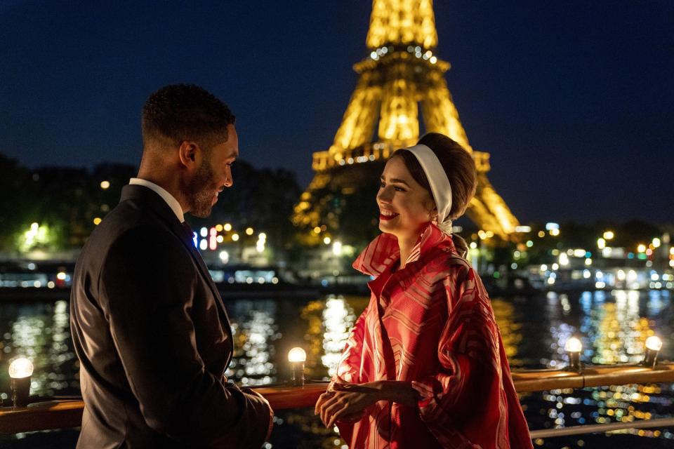 Still from "Emily in Paris" featuring Lucien Laviscount (L) and Lily (R) with the Eiffel Tower and the Seine River in the background at nighttime