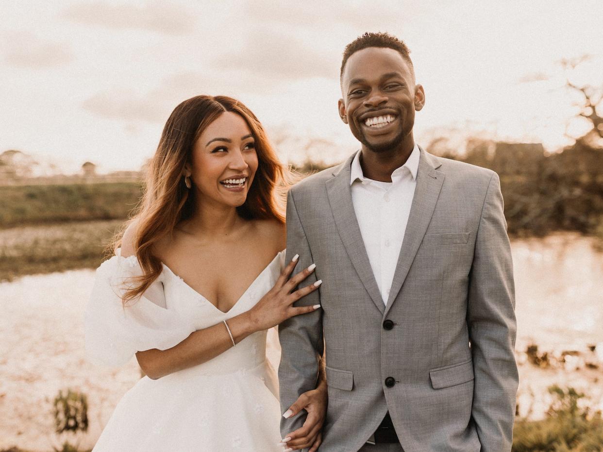 A bride looks at her groom and laughs as he smiles.They stand in front of a pond.