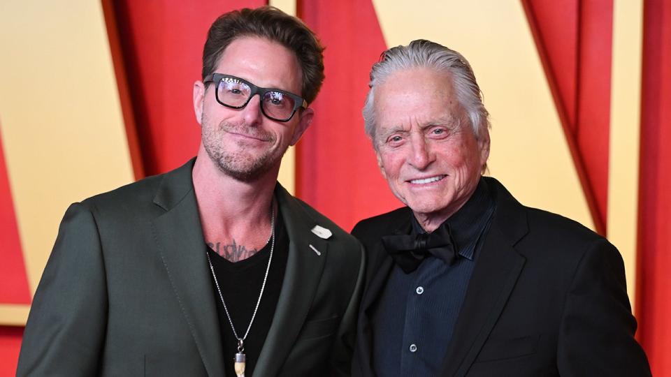 Cameron Douglas in a green suit and black framed glasses smiles next to dad Michael Douglas at the Vanity Fair Oscar party