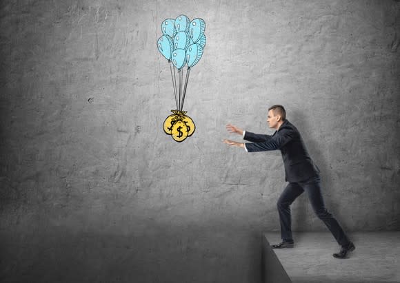 A businessman reaching for a bag of money suspended by balloons over a cliff.