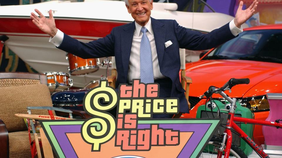 bob barker sits in a directors style chair and opens his arms wide, he is wearing a blue suit and tie and is surrounded by products including a speed boat, red car, drum kit, red bicycle, and snow sled, in front of him is a multicolored sign that says the price is right