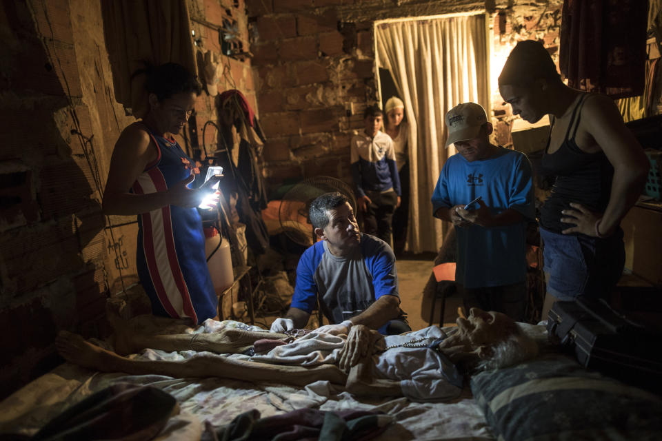 Community activist Carolina Leal uses a cell phone to light up the work area for Roberto Molero as he prepares the body of Teresa Jimenez, 91, after she died of natural causes in her home in Maracaibo, Venezuela, Nov. 17, 2019. Molero embalms bodies with no training other than seeing it done during a decade that he work as a driver at a funeral home, while Leal assumes the role of funeral director in her poor and often violent Maracaibo neighborhood, hoping to rid families of unnecessary misery she's seen too many times. (AP Photo/Rodrigo Abd)