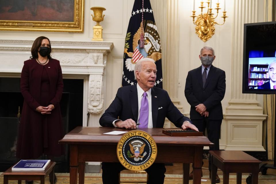 President Biden sits at a desk as Vice President Kamala Harris and Dr. Anthony Fauci stand behind him.