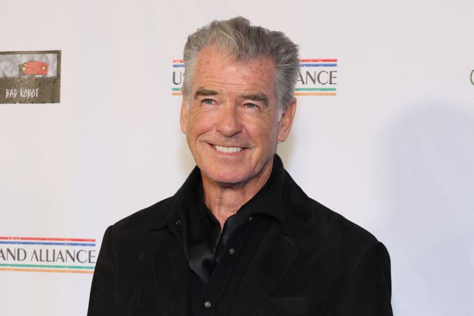 Pierce Brosnan stands smiling in a black shirt at a promotional event