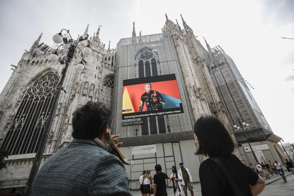 Pedestrians pass by a screen on the Duomo cathedral, showing a Moschino model during the Milan Digital Fashion Week, in Milan, Italy, Tuesday, July 14, 2020. Forty fashion houses are presenting previews of menswear looks for next spring and summer and pre-collections for women in digital formats, due to concerns generated by the COVID-19. (AP Photo/Luca Bruno)