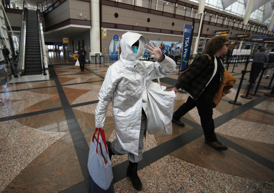 An unidentified traveler dressed in protective gear waves as she heads to the security checkpoint on the way to catching a flight out of Denver International Airport as passengers deal with the spread of the coronavirus, March 18, 2020, in Denver. (AP Photo/David Zalubowski)