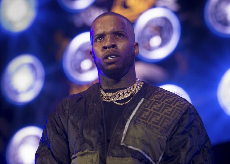 A rapper in a dark jacket and multiple gold chains looks slightly upward while onstage