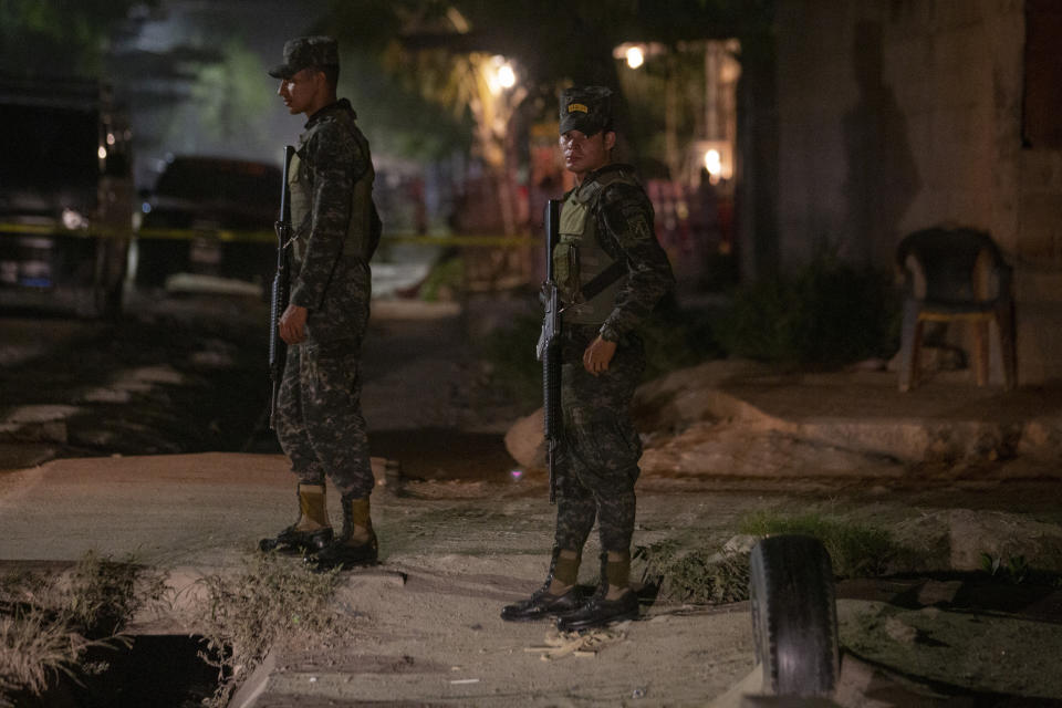 In this Nov. 30, 2019 photo, soldiers stand guard near a crime scene as forensic workers inspect a body in the Rivera Hernandez neighborhood of San Pedro Sula, Honduras. There are police stations in these neighborhoods, but everyone knows who is in charge. The gangs monitor the streets, the police patrols and rival gangs using a complex network of young boys who work in shifts around the clock and report anything suspicious. (AP Photo/Moises Castillo)