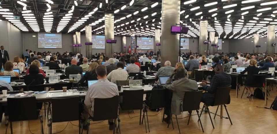 Shot from behind, a room full of people sitting at rows of desks with screens in the background.