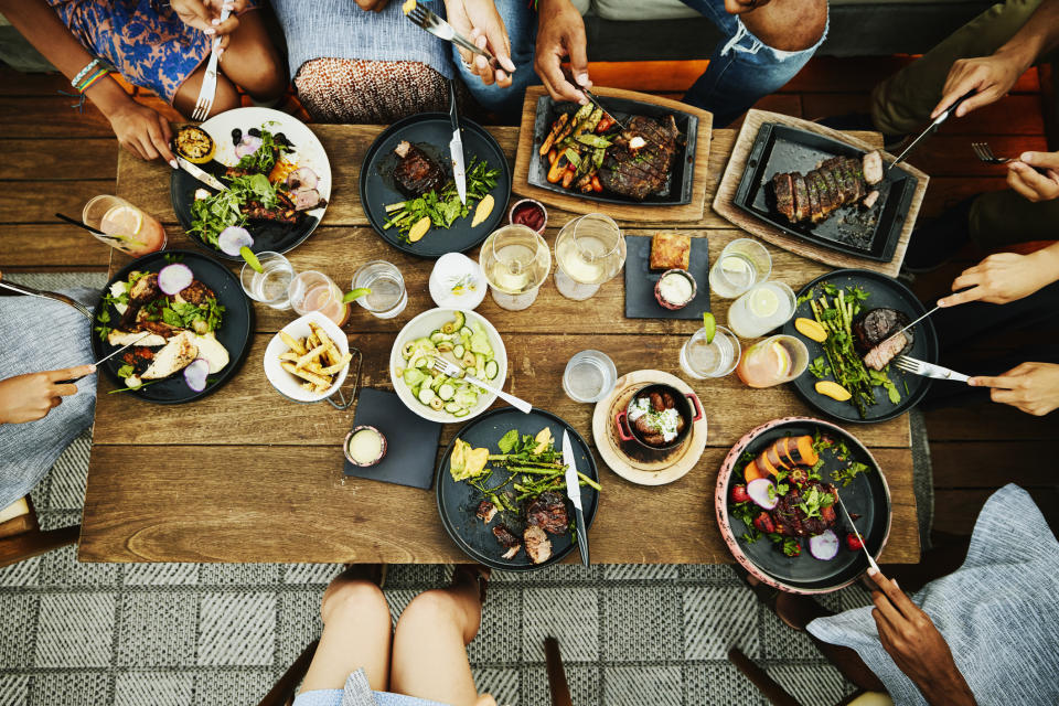 A group of people dining together at a table, with various dishes including steak, salad, and fries, and drinks on wooden table. Hands are holding utensils