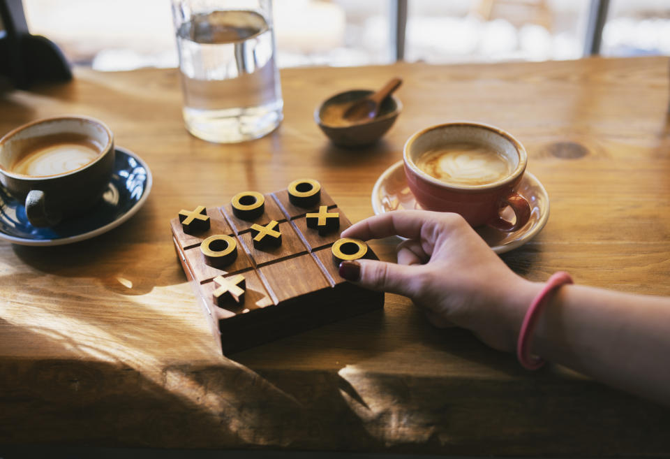 Hand of a person playing tic-tac-toe on a wooden board, with coffee cups and a water bottle on a wooden table