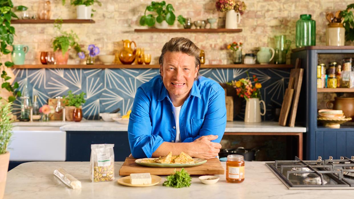  Jamie Oliver in a blue shirt leaning against a kitchen counter for Jamie's 5 Ingredient Meals. 