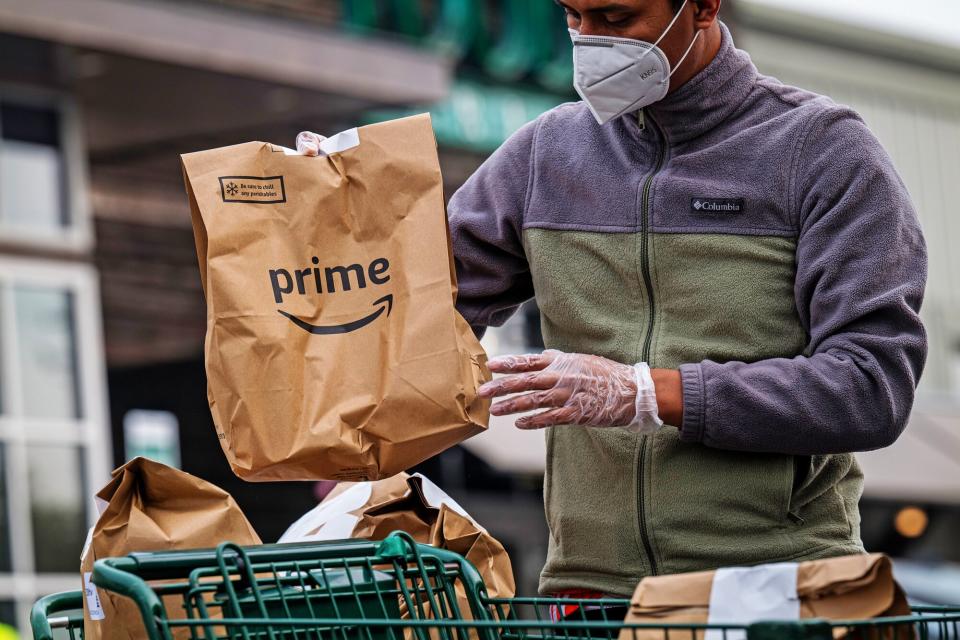 An independent contractor wearing a protective mask and gloves loads Amazon Prime grocery bags into a car outside a Whole Foods Market