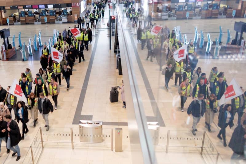 Aviation security staff demonstrate with flags in the departure hall in Terminal 1 at Berlin Brandenburg Airport. According to industry estimates, tens of thousands of passengers will once again be unable to travel as planned on Thursday due to warning strikes by aviation security staff at BER and elsewhere. The trade union Verdi has called further warning strikes by aviation security staff for this Friday. Carsten Koall/dpa