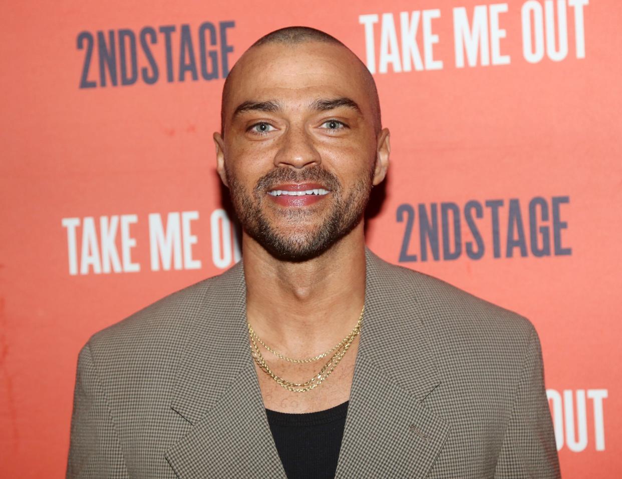 Jesse Williams in front of an orange backdrop in a brown suit jacket and black shirt