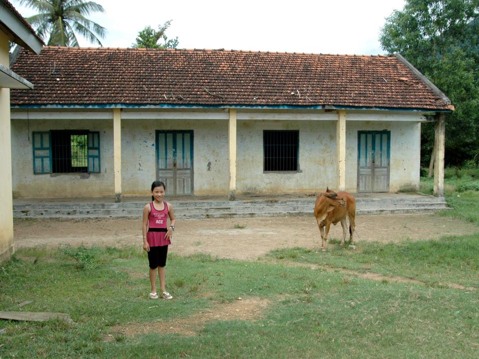Ha Nguyen pictured outside her old schoolhouse in Vietnam. The students drank a glass of milk every day supplied by the cow in the photograph.