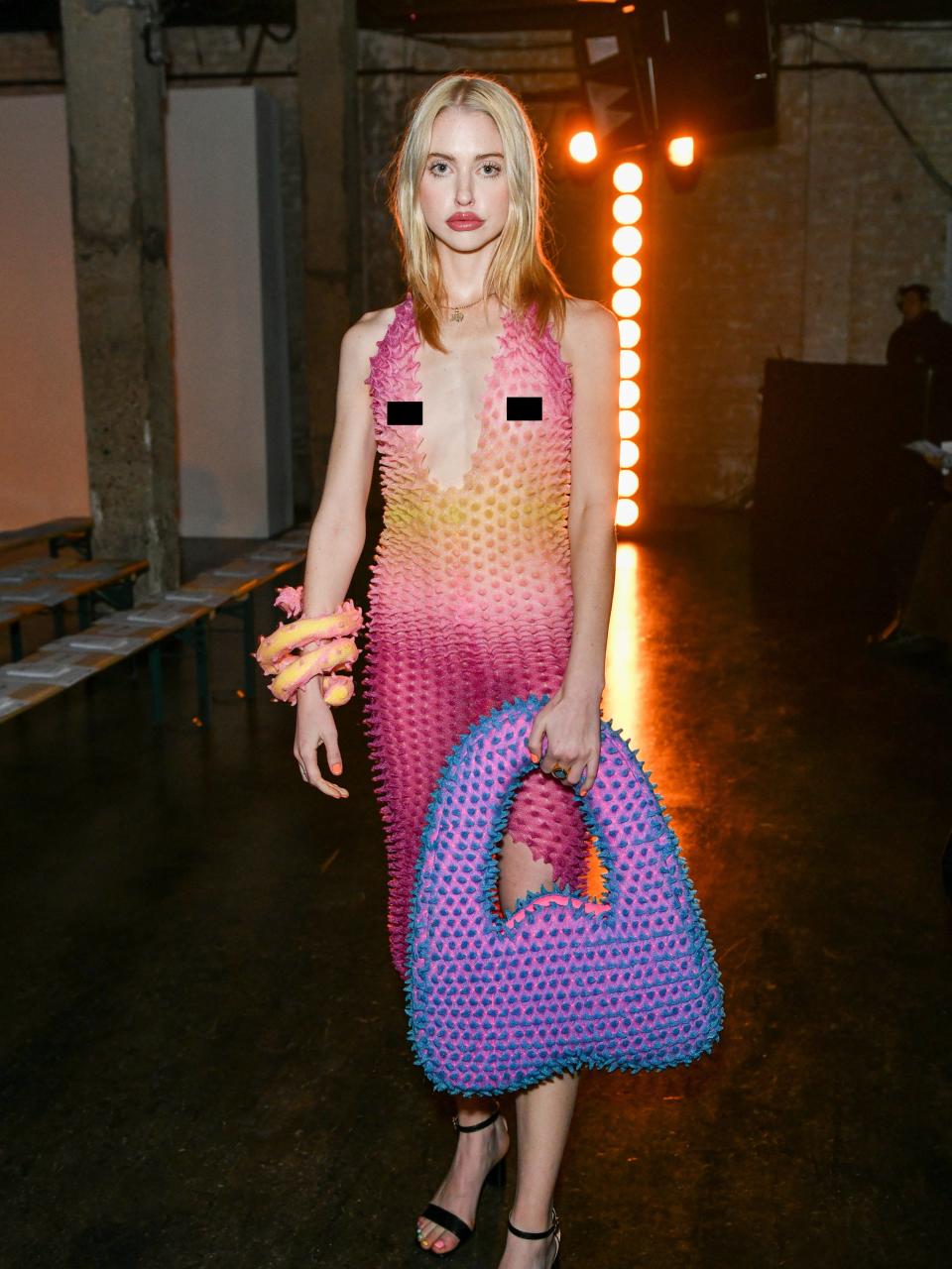 Chloe in a sheer pink and yellow gradient halter dress with raised dots. She has a matching oversized bangle wrapped around her best and an oversized purple and blue purse with matching raised dots.