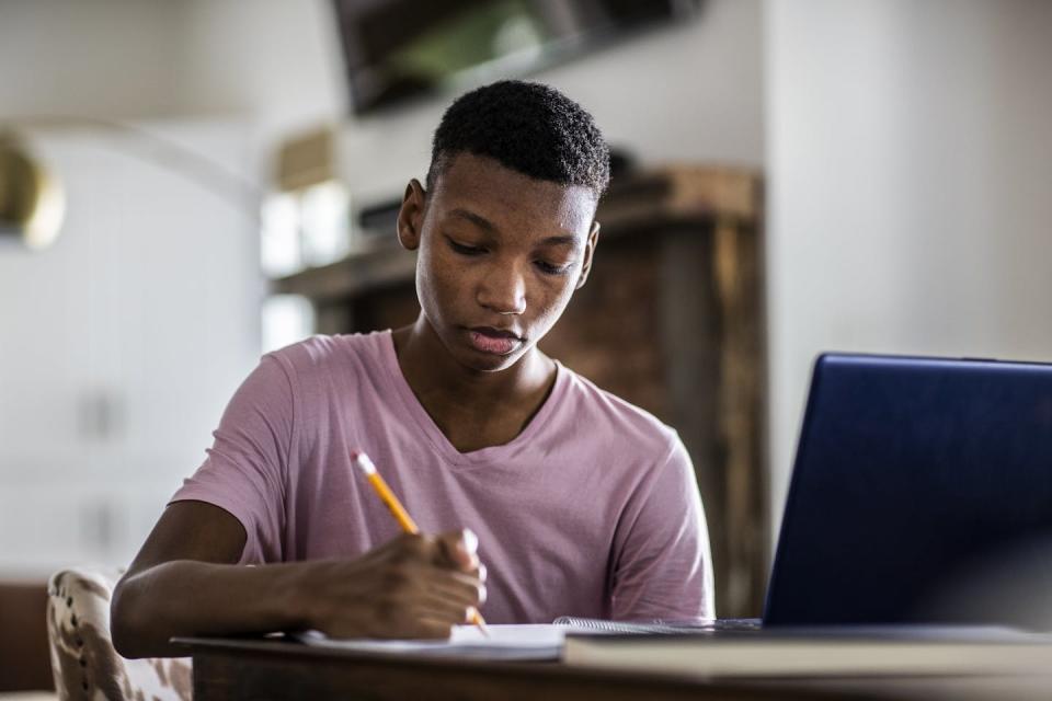 Writing on a notepad with his laptop nearby, a teenage boy does his homework.