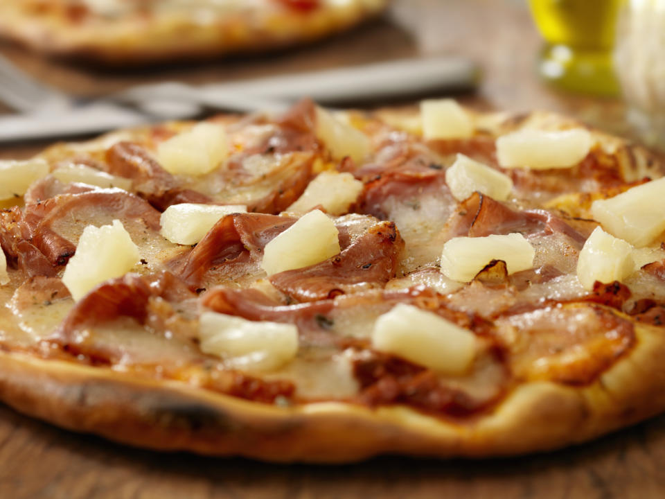 Pizza with ham and pineapple toppings on a wooden table. Another pizza and an olive oil bottle are slightly visible in the background