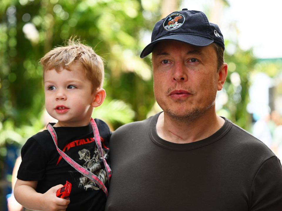 Elon Musk wears a baseball cap and holds his son in his arm