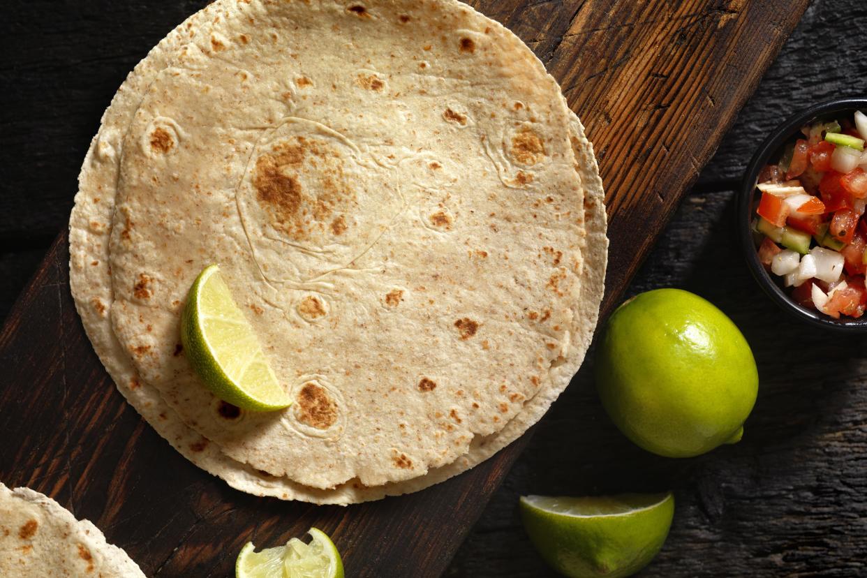 A pile of wheat flour tortillas with a slice of lime on a dark wooden cutting board surrounded by limes, lime slices and salsa