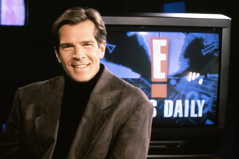 TV host in studio with 'E! News Daily' on screen, wearing a suit jacket, smiling at the camera