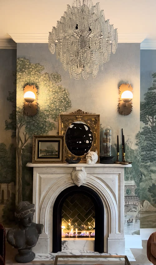 An antique Victorian marble fireplace mantel set against scenic wallpaper installed by Mantel House LLC, decorated with mirrors, art and candlesticks.