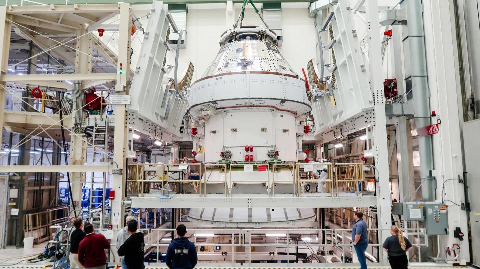 a white-and-silver cone-shaped spacecraft is held aloft by a crane inside a room full of machinery and scientists