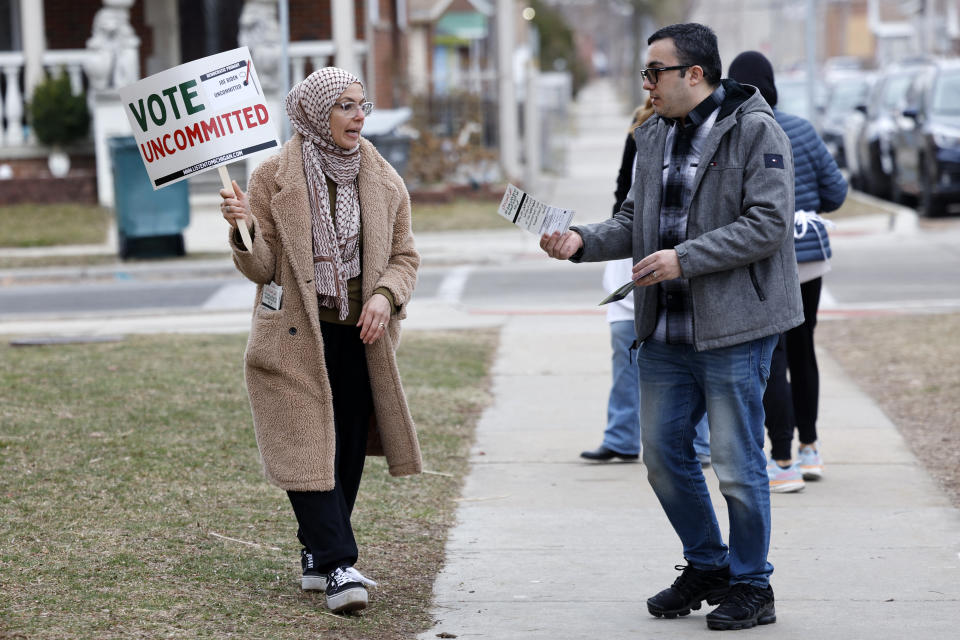 A woman wearing a headscarf and winter coat stands along a sidewalk near several other people and holds a sign reading: Vote uncommitted.