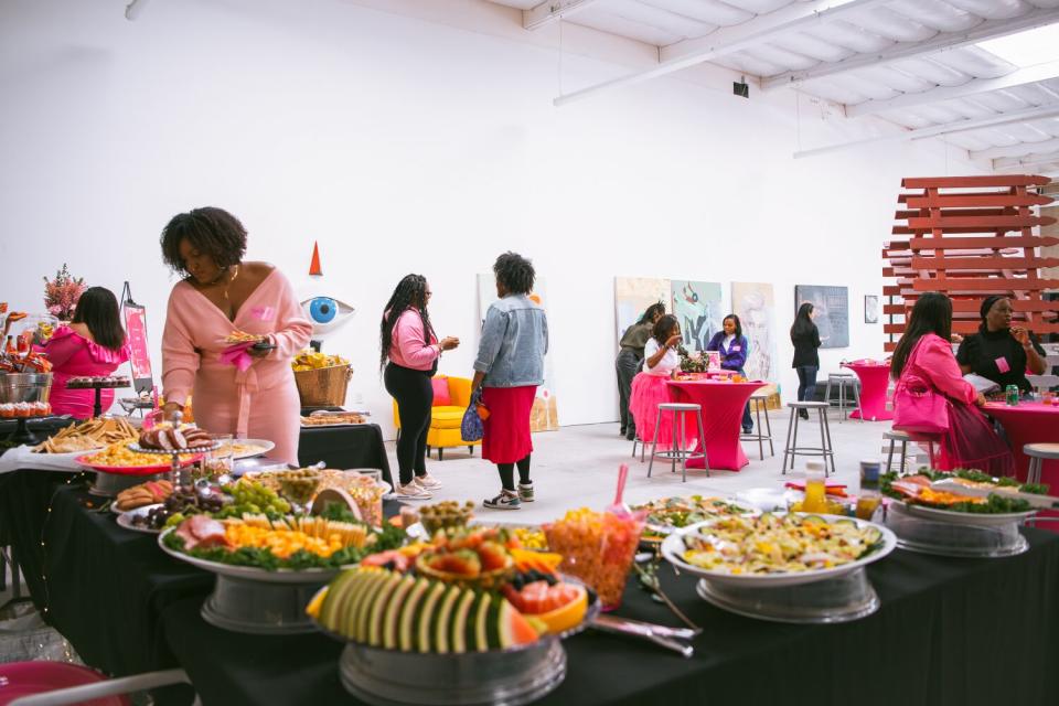 A group of women scattered around a room, sitting at tables and serving themselves from a buffet in the foreground.