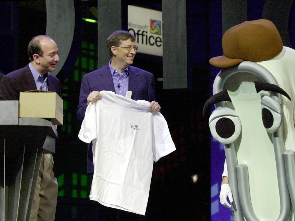 Former Microsoft CEO Bill Gates and former Amazon CEO Jeff Bezos hand a white t-shirt to someone dressed in a cartoon paperclip costume.